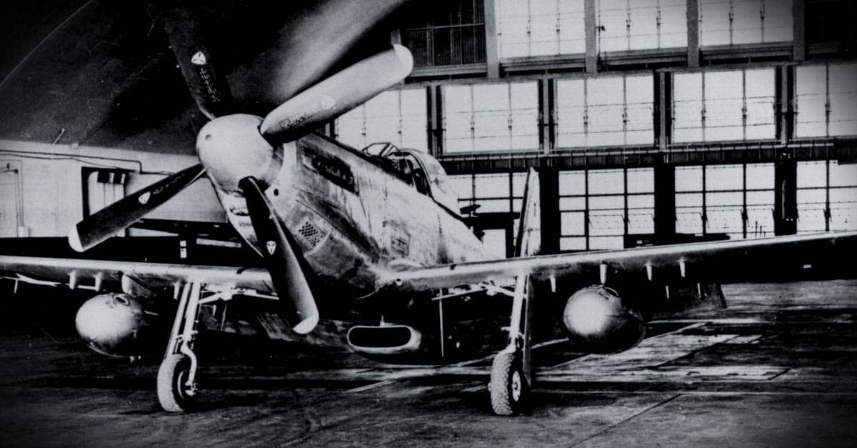 A P-51D Mustang shown inside a maintenance hangar with extended range drop tanks and hard points
